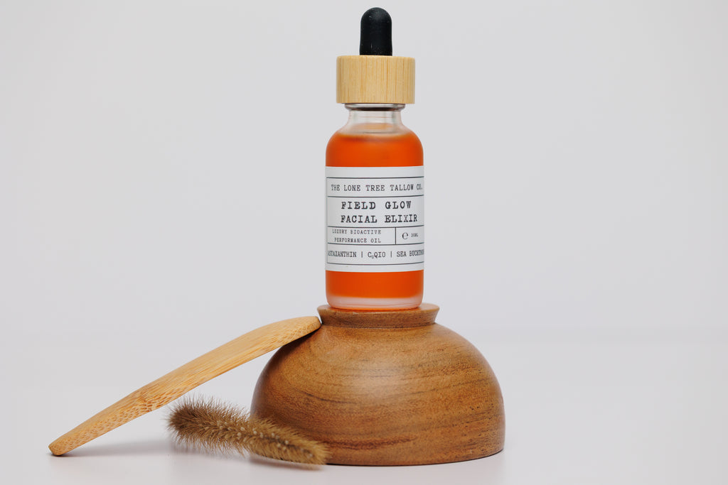 1 oz dropper bottle of Field Glow Luxury Facial Oil Elixir sitting on an upside down wooden bowl next to a wooden scoop and a sprig of grain, all  on a white background. Field glow contains astaxanthin, sea buckthorn, CoQ10, rosehip oils and can be compared to luxury oils like Oliver Midy and Vintner's daughter for a fraction of the price. 