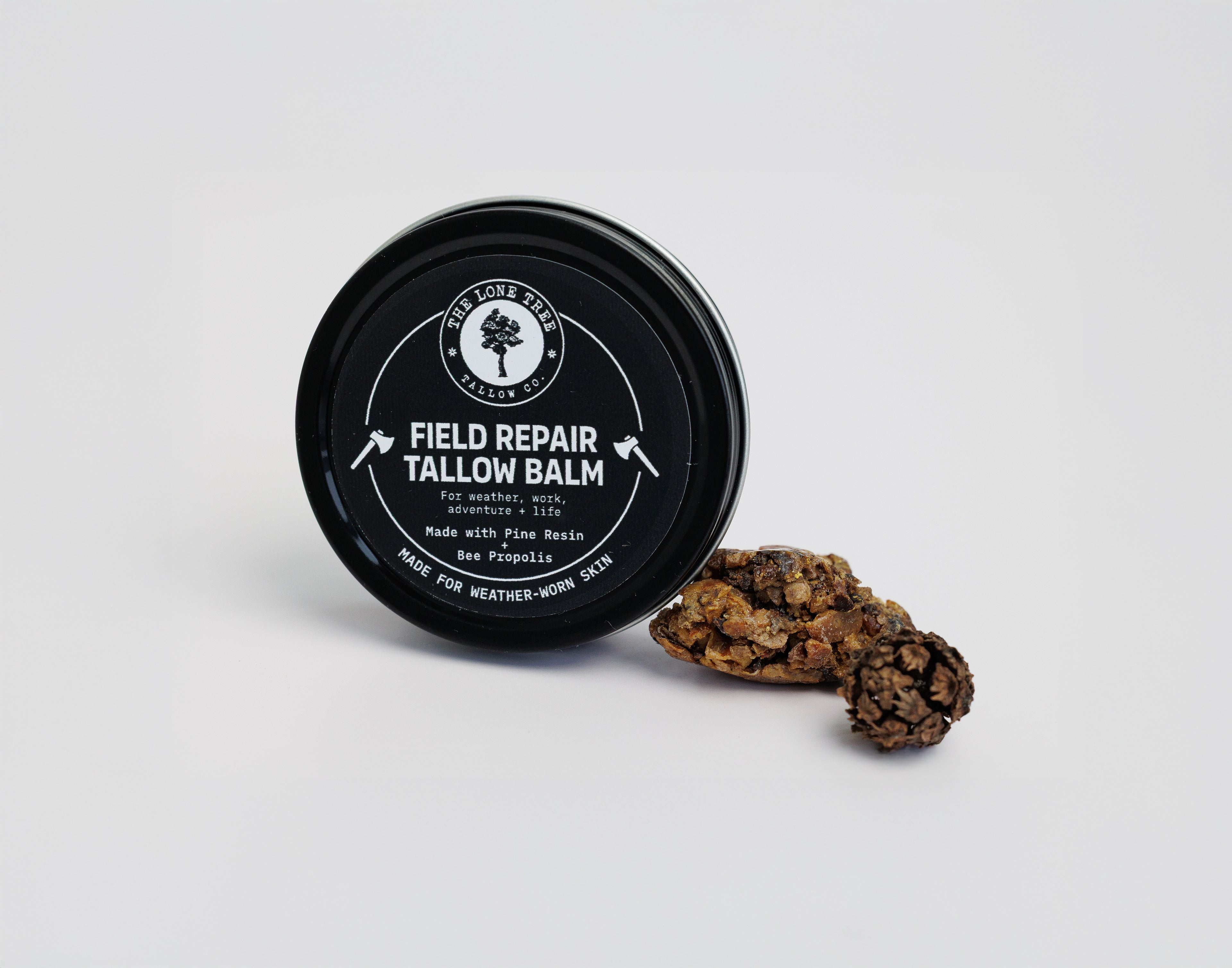Field Repair Beef Tallow Balm with bee propolis, pine resin, shea butter, calendula infused sunflower oil heals dry hands, cracked skin and is hand crafted on the prairie. shown here next to pine resin in a 2 oz black tin. 