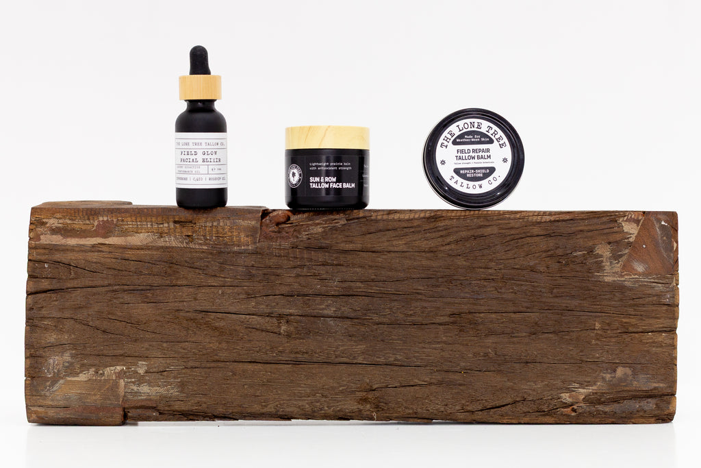 The Men's Skincare Kit: Black bottle of the Field Glow Facial Oil Elixir with Astanxanthin and CoQ10 sitting next to the Sun & Row Antioxidant Tallow Face Balm and a black tin of the Field Repair Tallow Blam with Bee Propolis and Pine Resin. All products are sitting on a rustic piece of wood with a white background. 